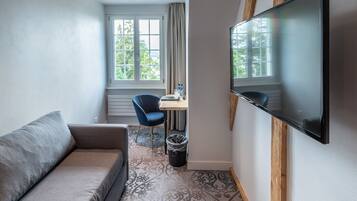 A compact hotel room with a sofa, a small desk with a chair, a television mounted on the wall, and a window with curtains.