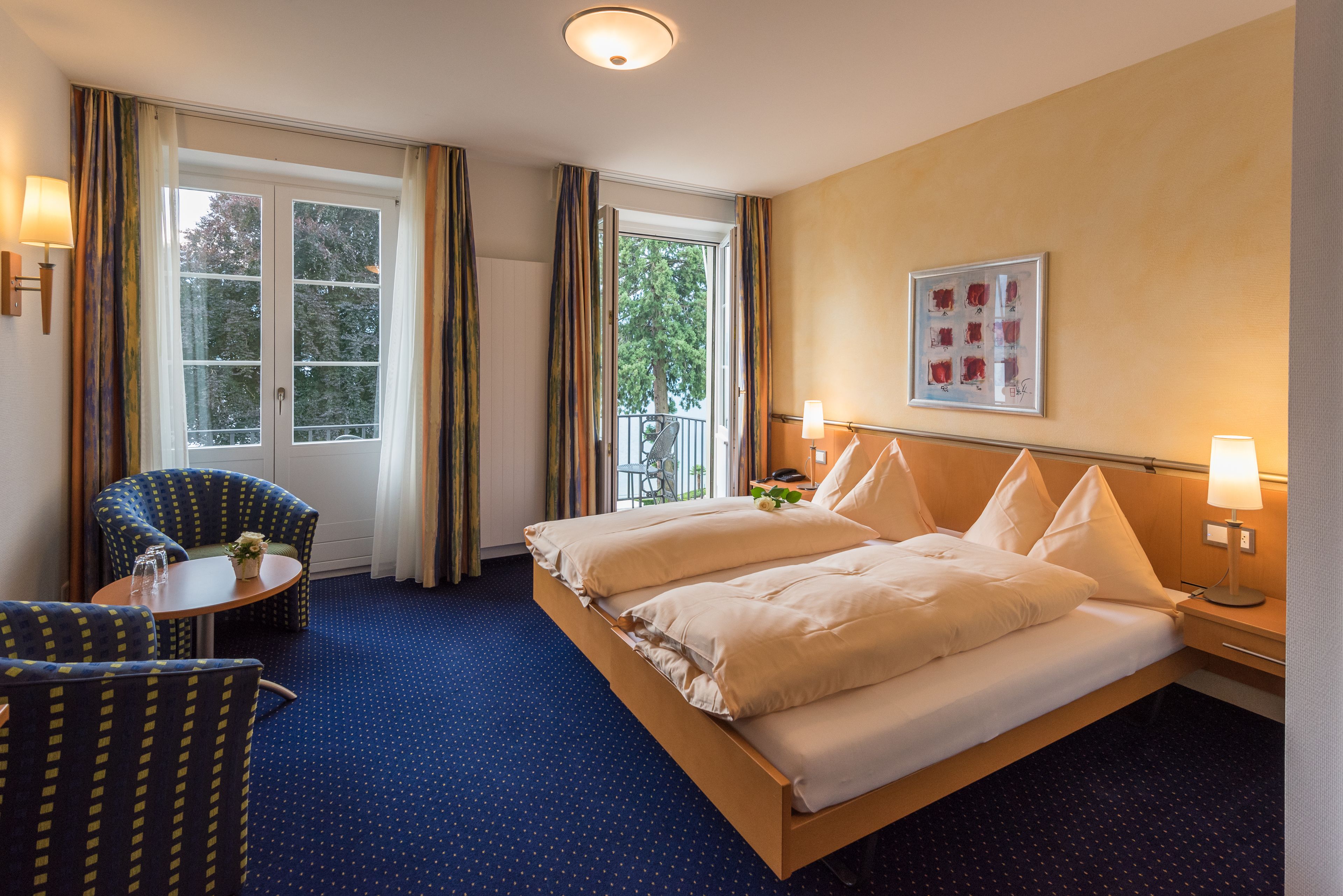 Une chambre d’hôtel avec un grand lit, deux chaises, une petite table et une fenêtre avec des rideaux.
