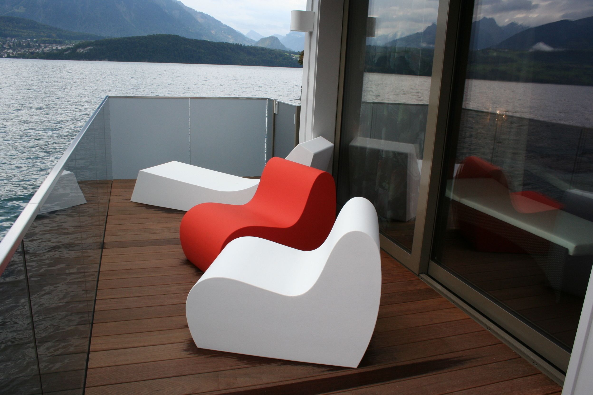 A modern deck with two white and one red lounge chair, overlooking a calm lake with mountains in the distance.