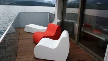 A modern deck with two white and one red lounge chair, overlooking a calm lake with mountains in the distance.