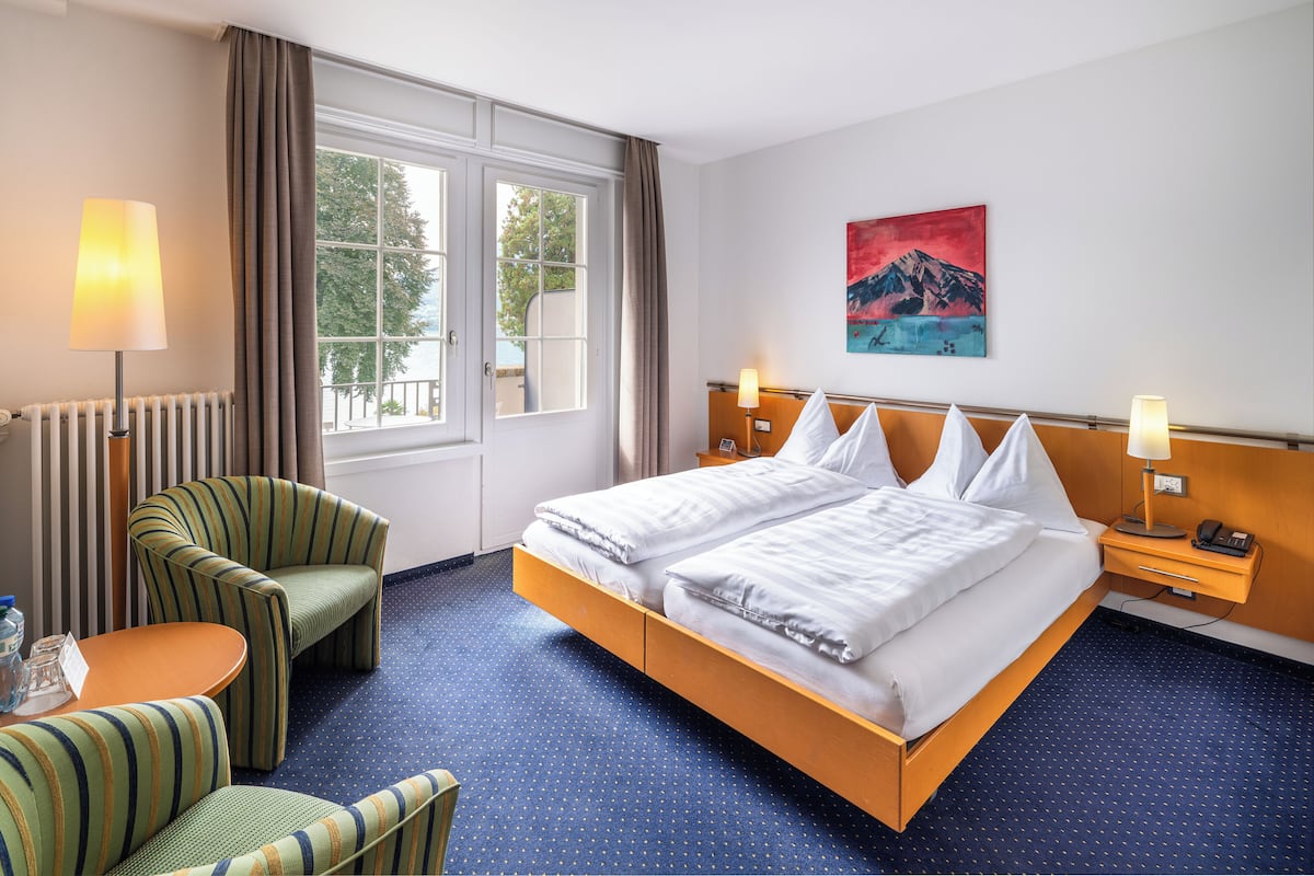 Una habitación de hotel con una cama grande, dos mesitas de noche con lámparas, una silla, una pequeña mesa y una ventana con cortinas.