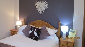 A neatly made bed with a heart-shaped wall decoration, two bedside lamps, and a nightstand with a lamp and a sign. - Broomlea (Inverness)
