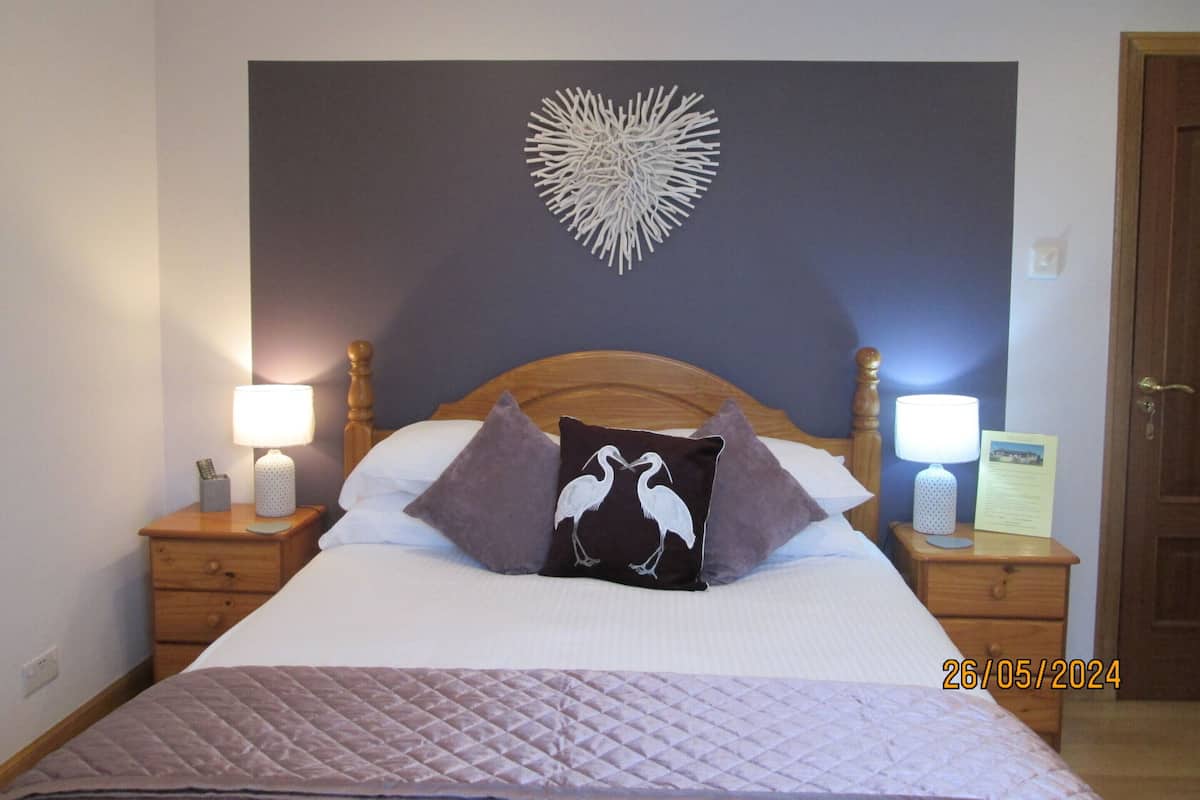 A neatly made bed with a quilted cover, two bedside lamps, a wooden headboard, and a wall decoration.