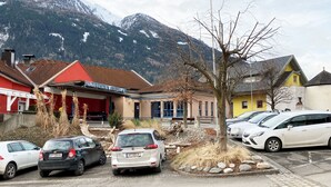 Una àrea de pàrquing amb diversos cotxes, un edifici amb un rètol que diu "KULTURZENTRUM OEHELVLIAC" i un paisatge de muntanyes al fons.