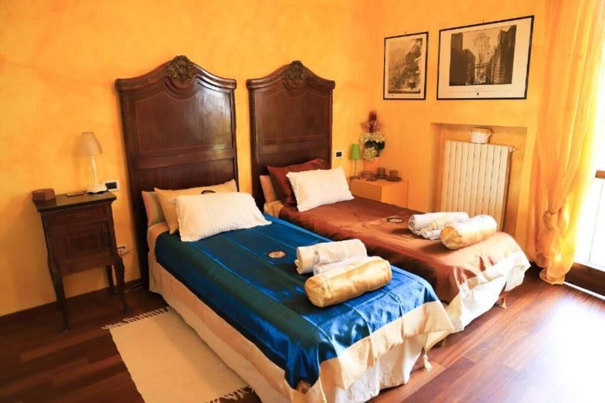 Une chambre d’hôtel avec deux lits, des têtes de lit en bois, une table de chevet avec une lampe, un radiateur et deux tableaux encadrés au mur.