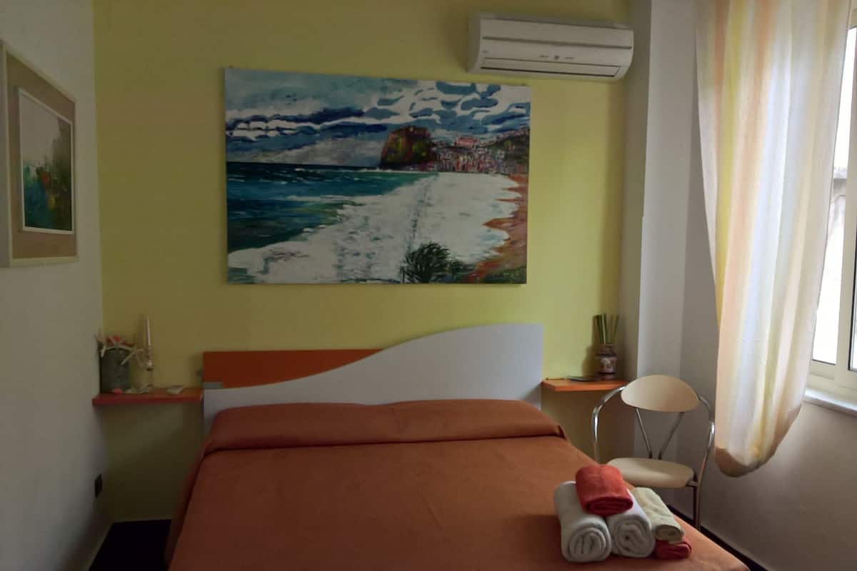 Habitación de hotel con cama, un cuadro de una escena de playa, una ventana con cortinas y una mesita con lámpara.