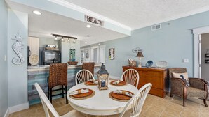 A dining area with a white table set for four, surrounded by wicker chairs. A kitchen area is visible in the background with a refrigerator and a sink.