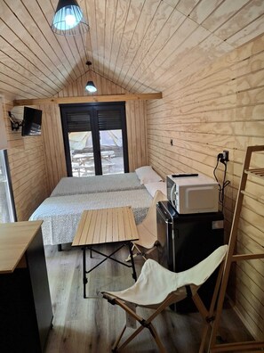 Eine kompakte Holzhütte mit einem Bett, einem kleinen Tisch, einem Klappstuhl, einer Mikrowelle und einem an der Wand befestigten Fernseher.