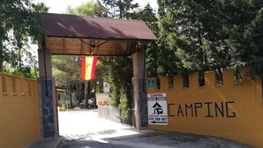 En grind markerar ingången till ett campingområde med ett träbeklätt tak, en gul vägg med en skylt som visar att det är ett campingområde, och en stenkiosk.