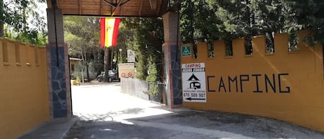 Una entrada cerrada a un área de camping con un toldo de madera, una pared amarilla con un letrero de camping y un pilar de piedra.