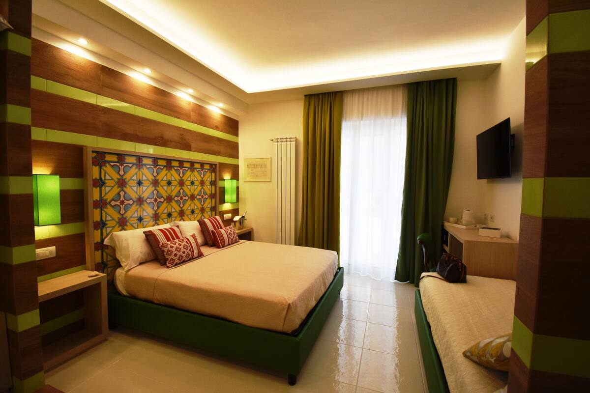 Een hotelkamer met een groot bed, een nachtkastje, een lamp, een aan de muur bevestigde televisie en een raam met gordijnen.