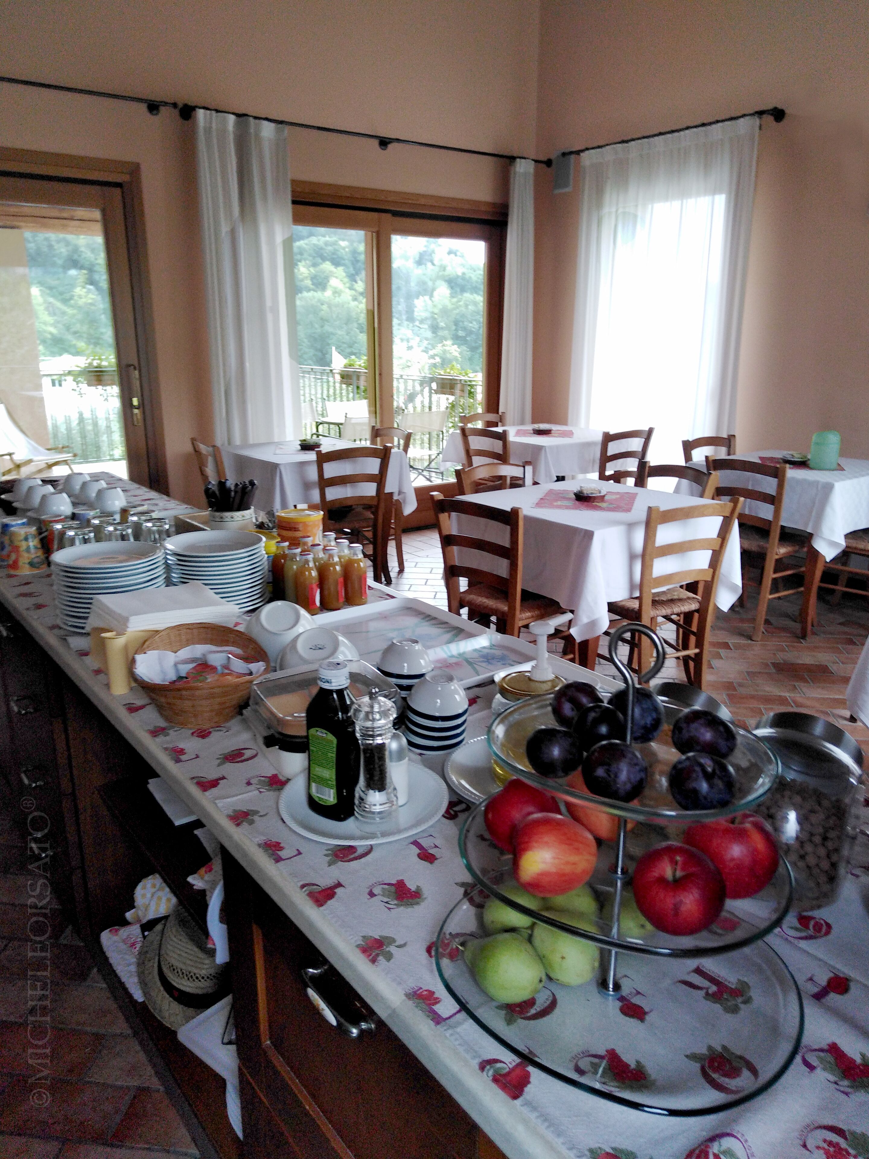 A breakfast buffet with various fruits, condiments, and dishes on a table in a bright room with windows.