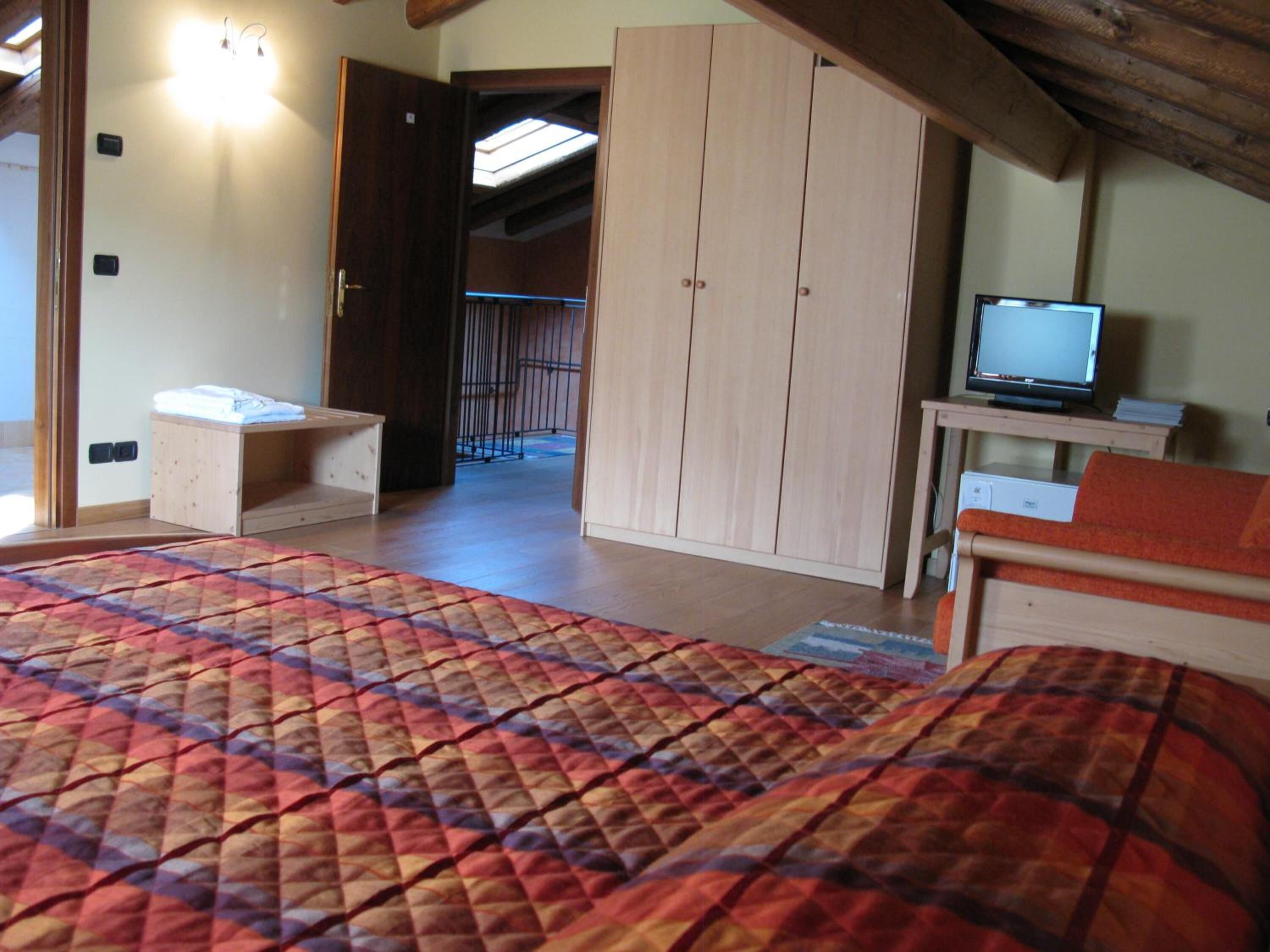 A bedroom with a bed, a wooden wardrobe, a television on a small table, and a bathroom visible through an open door.