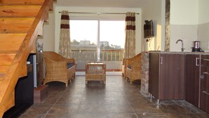 A spacious living area with a wooden staircase, wicker furniture, a kitchen area, and a balcony with curtains. - Cottages at Village (Nainital)