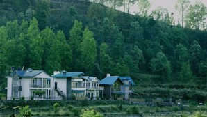 A row of modern two-story houses with a hillside backdrop, surrounded by lush greenery. - Cottages at Village (Nainital)