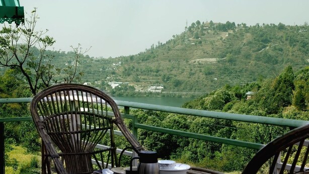A wicker chair with a metal cup and saucer, a coiled rope, and a metal pot on a balcony railing overlooking a green valley with buildings in the distance. - Cottages at Village (Nainital)