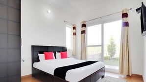 A modern bedroom with a large bed, a dark headboard, and a view of the outdoors through a sliding glass door. - Cottages at Village (Nainital)