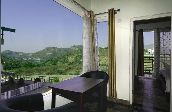 A balcony with a table and chairs, a view of green hills, and a door leading to a covered area. - Cottages at Village (Nainital)