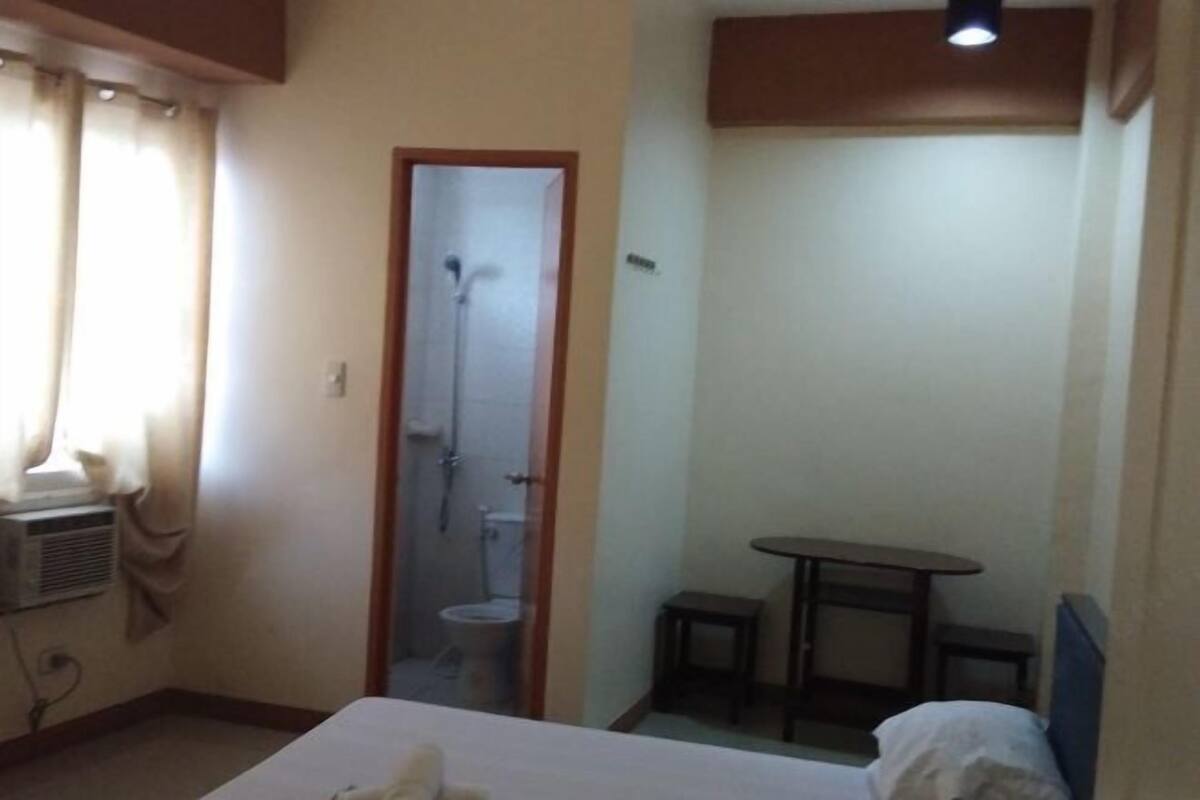 Habitación de hotel con cama, una mesita, una silla, zona de ducha y equipo de aire acondicionado.