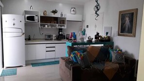 A compact kitchen with white cabinets, a microwave, and a coffee maker. A teal-colored table is in the center with a brown couch adorned with colorful pillows. - Apartamento Cantinho do Céu (Cachoeira Paulista)