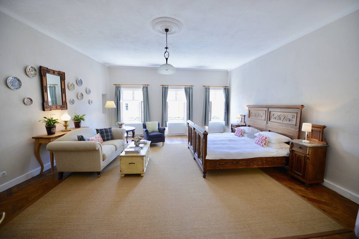 A spacious bedroom with a large bed, a sitting area with a sofa and armchair, a small table with a lamp, and a window with curtains.