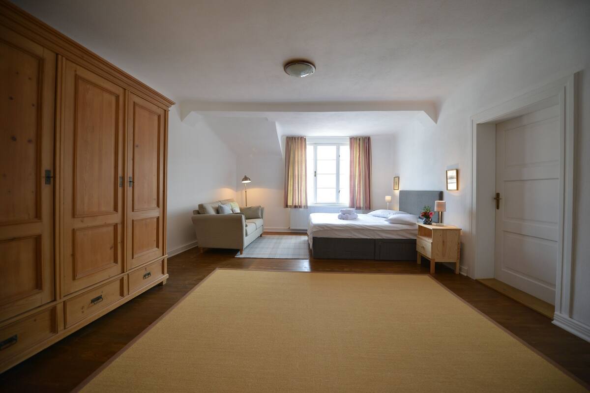A bedroom with a large wardrobe, a bed, a bedside table, and a window with curtains.