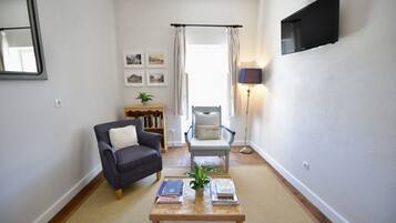 Un salon avec un canapé, un fauteuil, une table basse avec des livres, un téléviseur fixé au mur et une bibliothèque en arrière-plan.