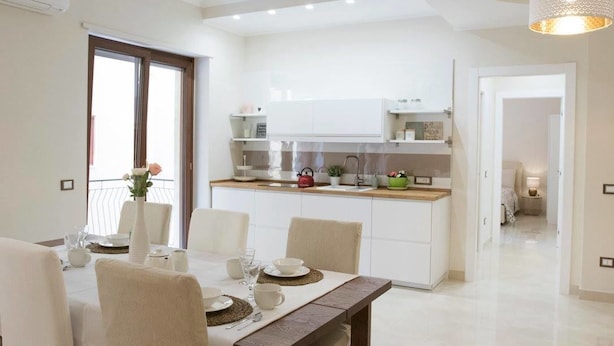 A modern kitchen with white cabinets, a dining area with a table set for four, and a view of the balcony through the window. - HomeAway B&B (Salerno)