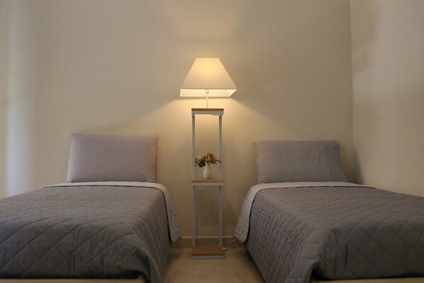 A room with two beds, a bedside table with a lamp, and a small vase with flowers.