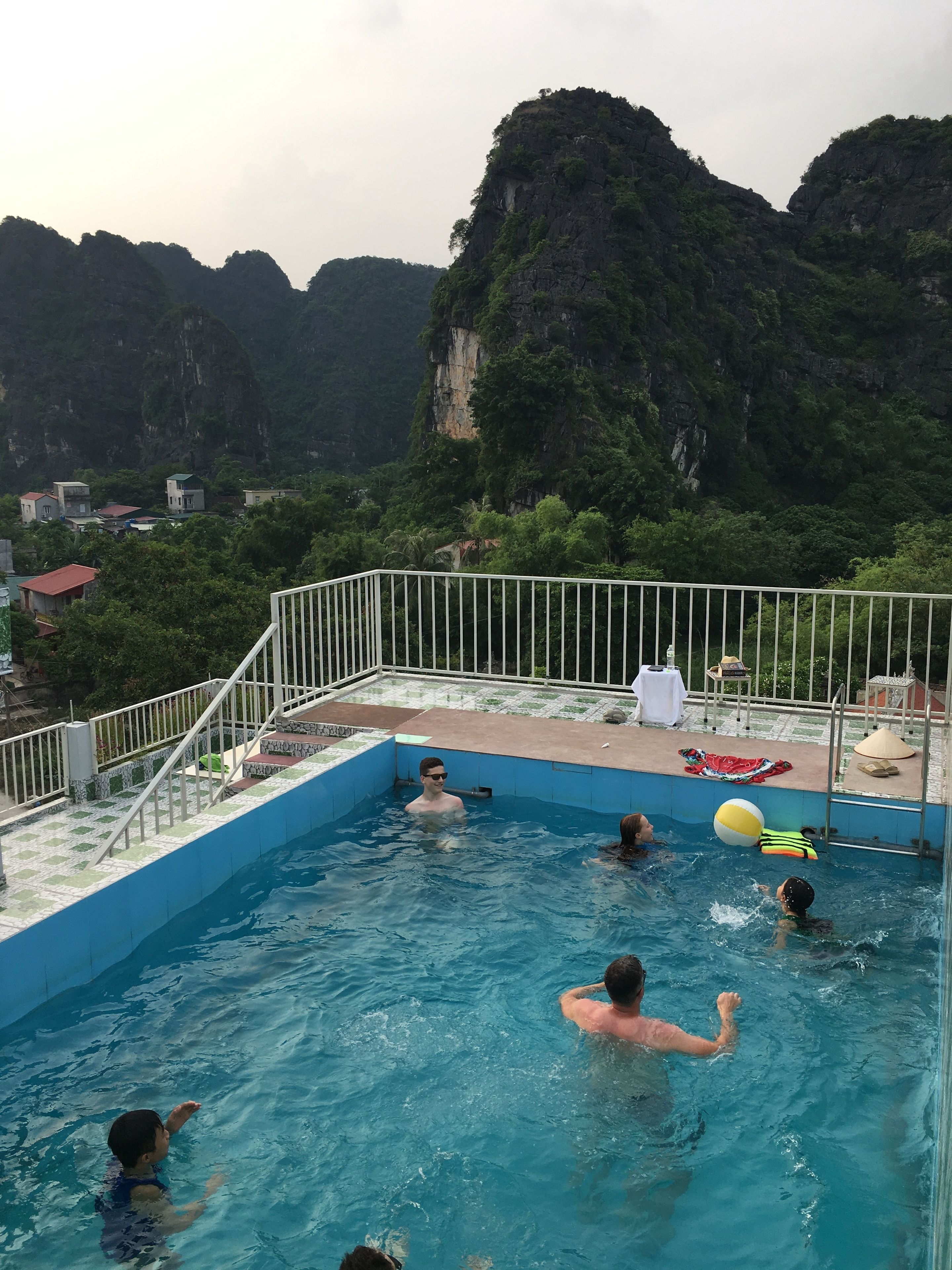 Una piscina con gente disfrutando del agua, rodeada por una barandilla y vistas a un paisaje montañoso.