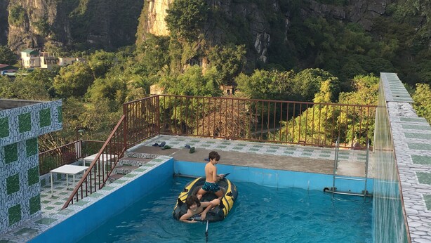 A swimming pool with two people on inflatable rings, surrounded by a tiled deck and a view of a mountainous landscape. - Limestone Boutique Hotel (Ninh Binh)