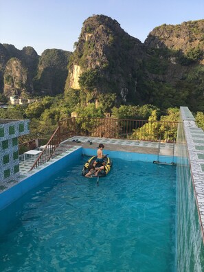 A swimming pool with two people on inflatable rings, surrounded by a tiled deck and a view of a mountainous landscape. - Limestone Boutique Hotel (Hoa Lu)