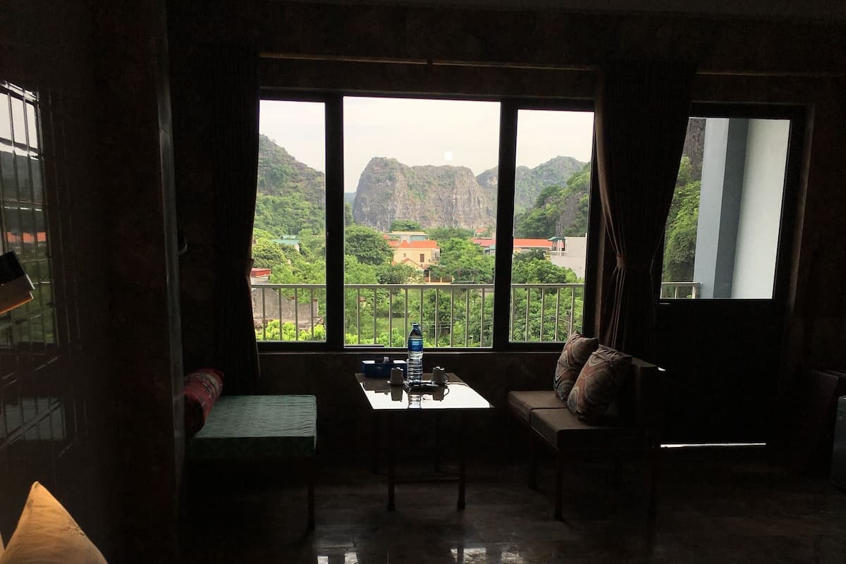 Una habitación con cama, una mesa y una botella de agua, y vistas a zonas verdes y montañas a través de amplios ventanales.