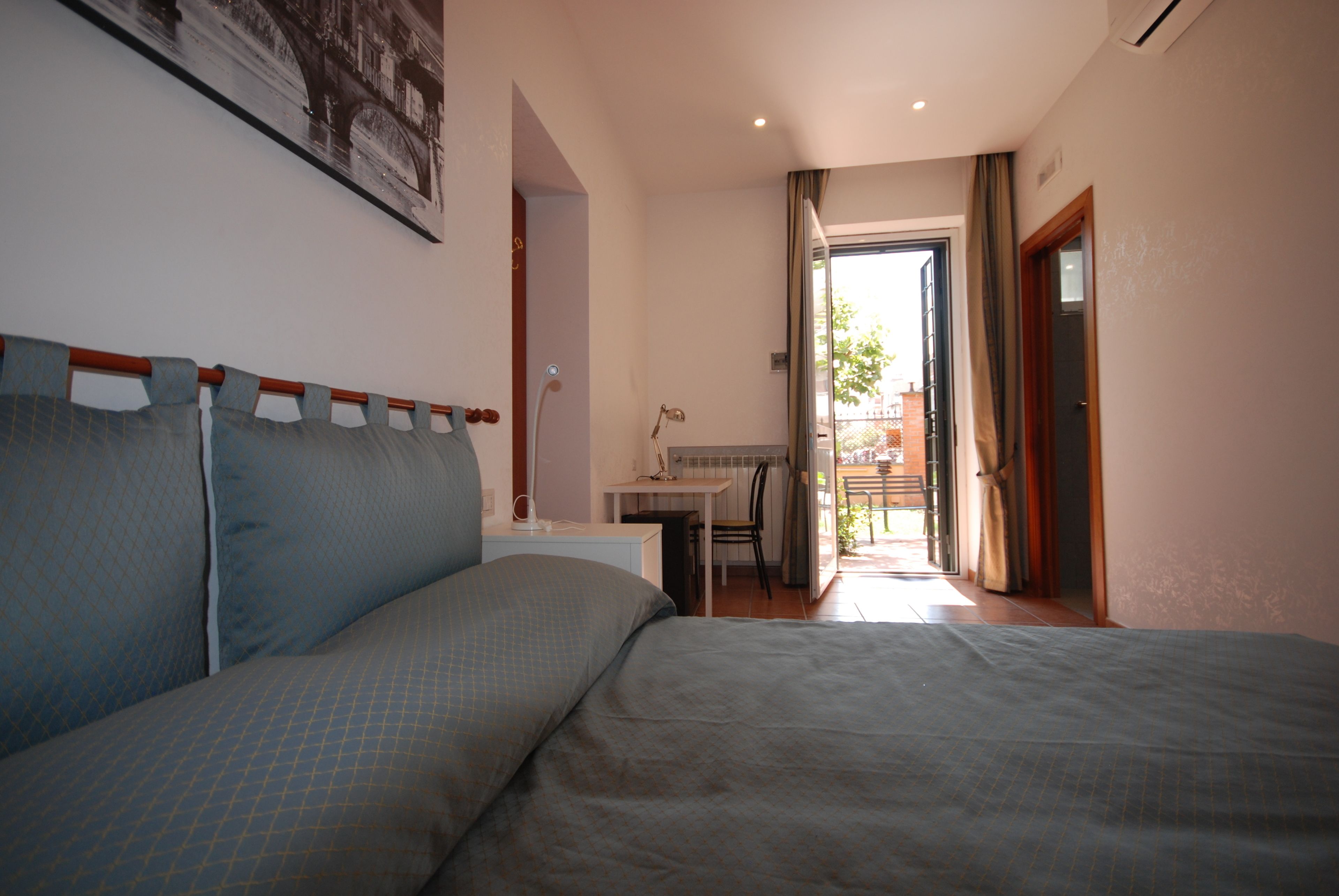 Habitación de hotel con cama, escritorio y silla. Se ve una ventana con cortinas y una puerta que da a un espacio al aire libre.