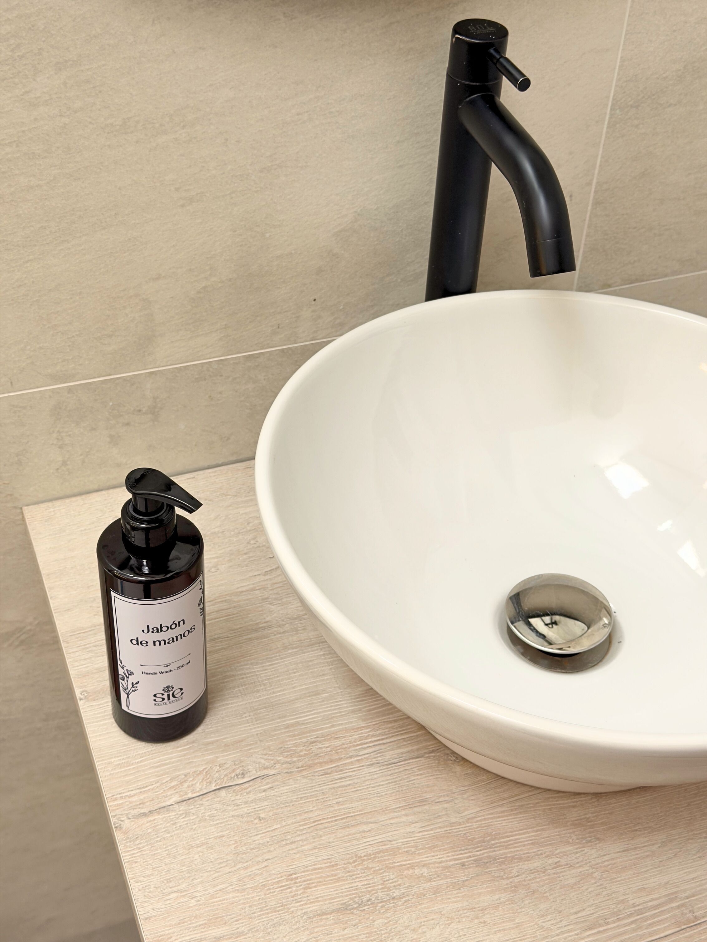 Un baño con lavamanos blanco, grifo negro y una botella de jabón para manos sobre una superficie de madera.