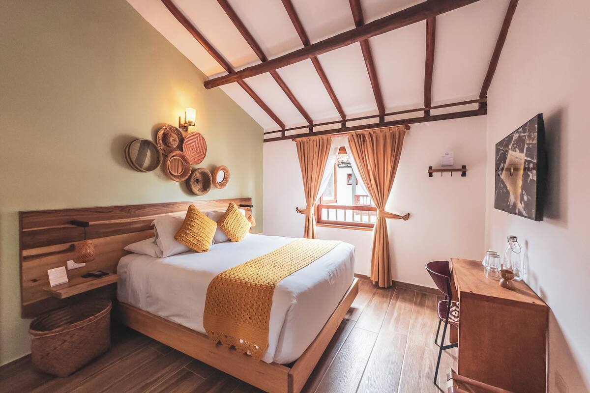 A bedroom with a wooden bed, a nightstand, a desk, a woven throw blanket, and a view of the outside.