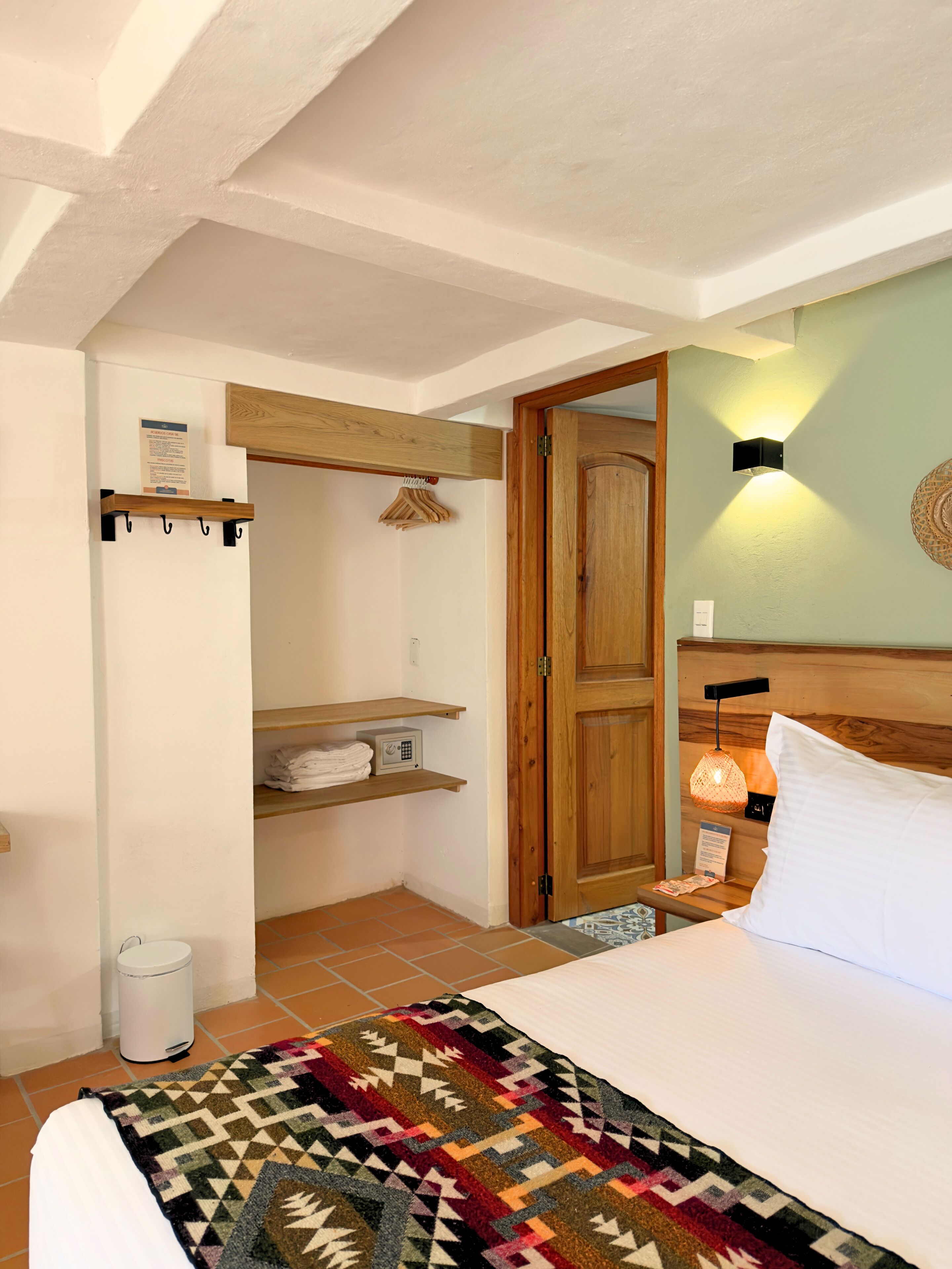 Un dormitorio con cama, mesita de noche, lámpara de pared, puerta de madera, estante con toallas dobladas y perchero.