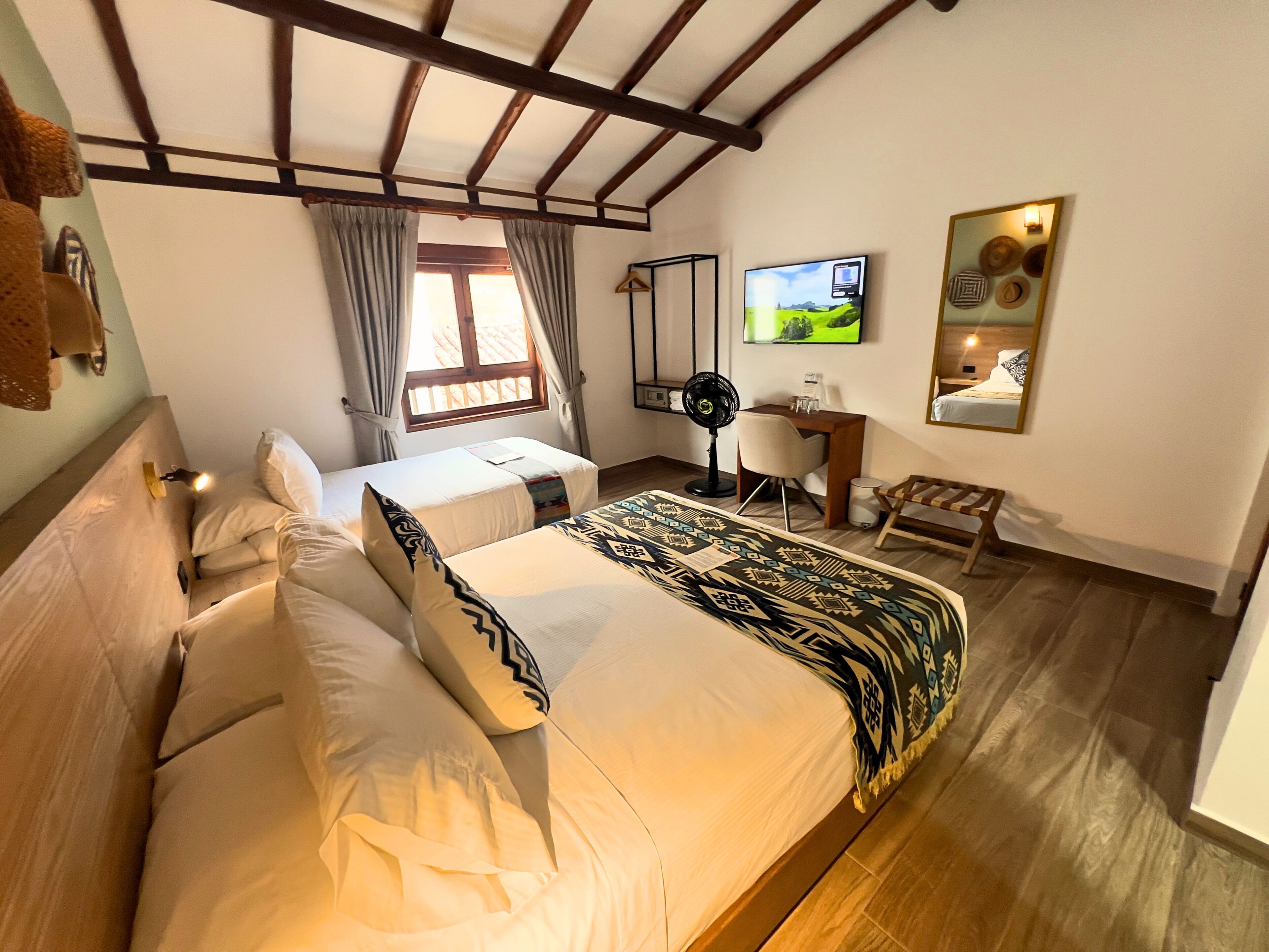Un dormitorio con una cama grande, un escritorio, una silla, un ventilador de pie y un televisor en la pared.
