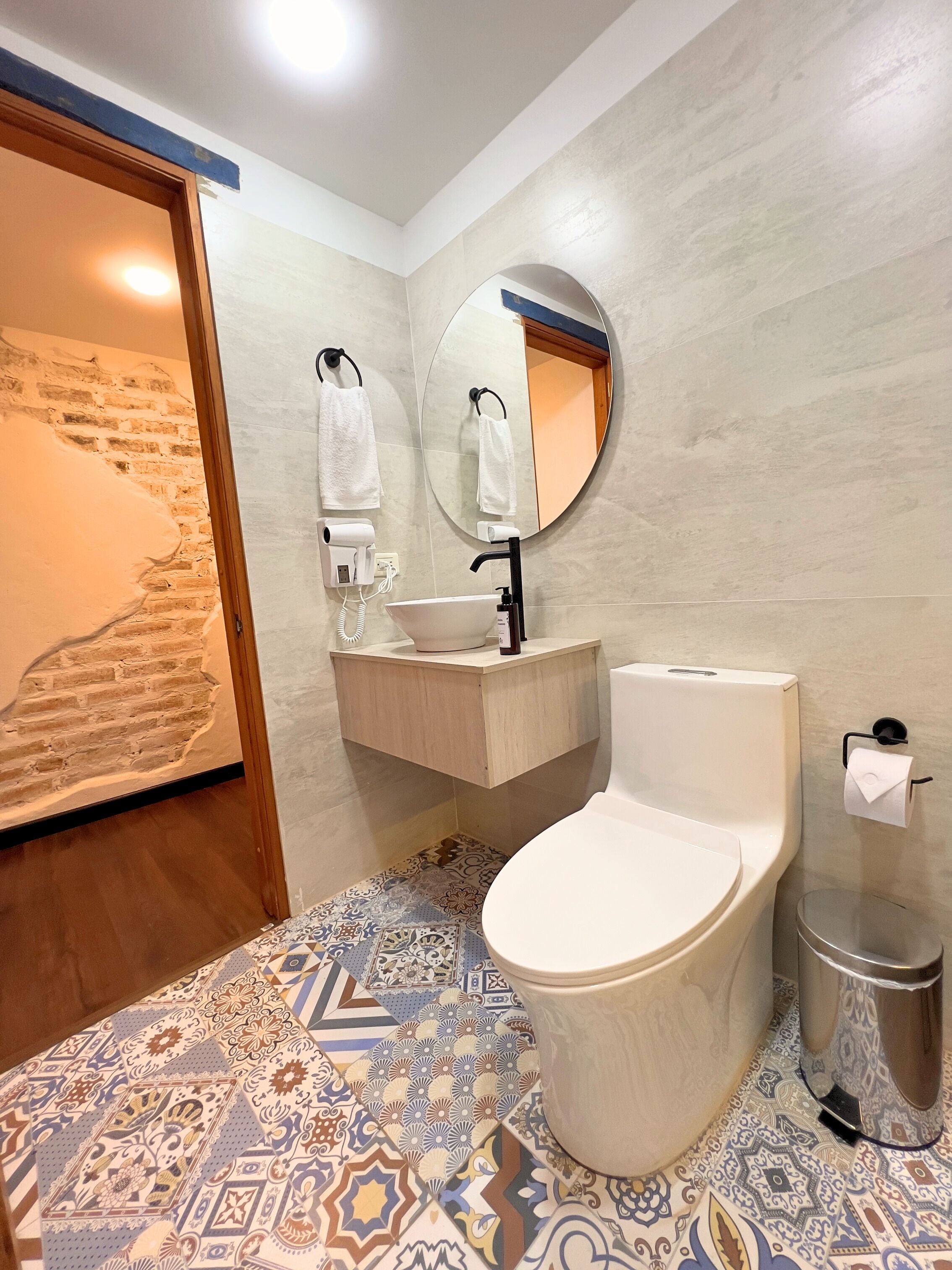 A bathroom with a toilet, sink, and mirror. There is a towel rack with towels, a trash can, and a wall-mounted hairdryer. The floor has patterned tiles.