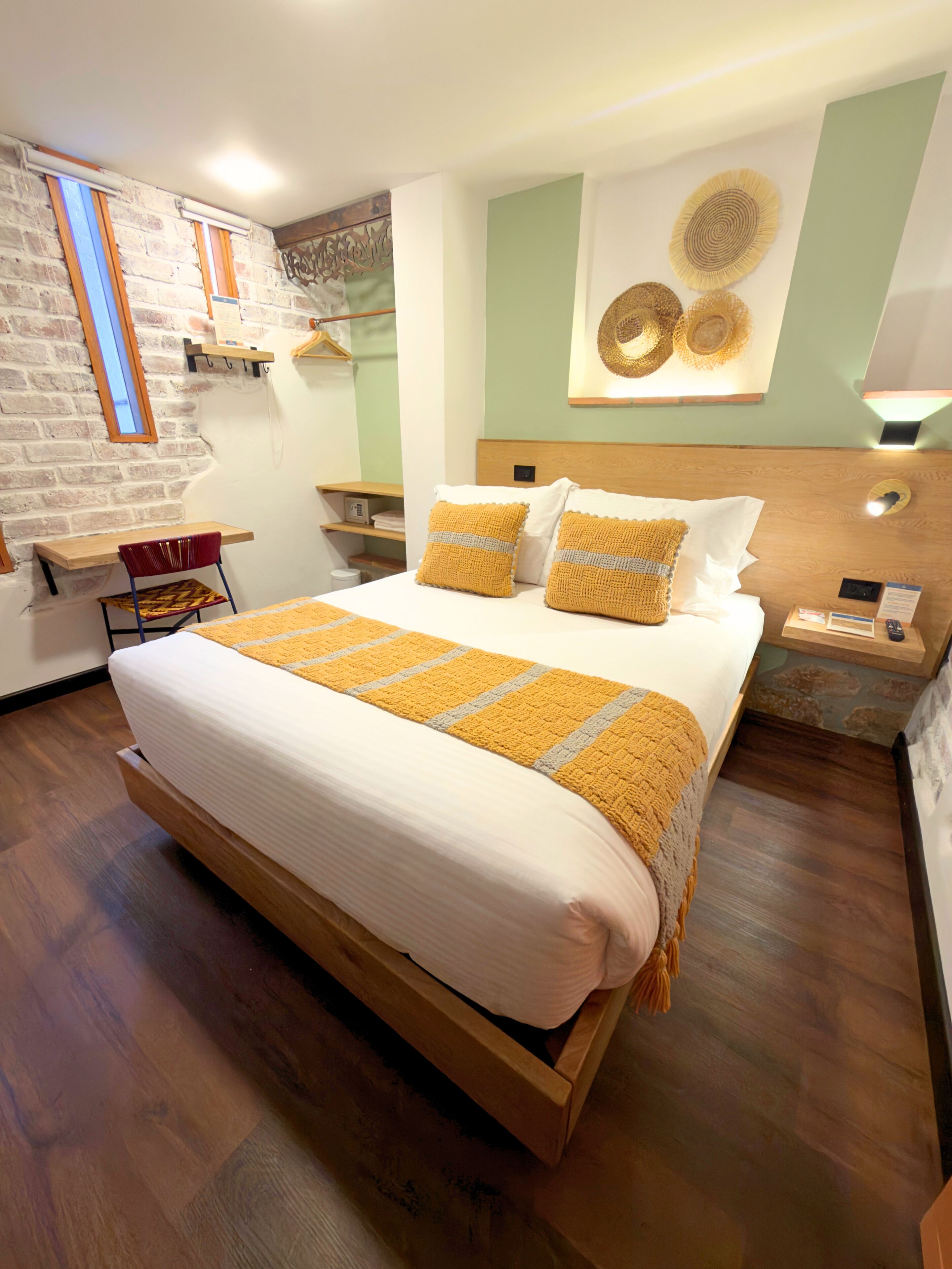 A hotel room with a large bed, wooden headboard, a small desk, and a brick wall with a window.