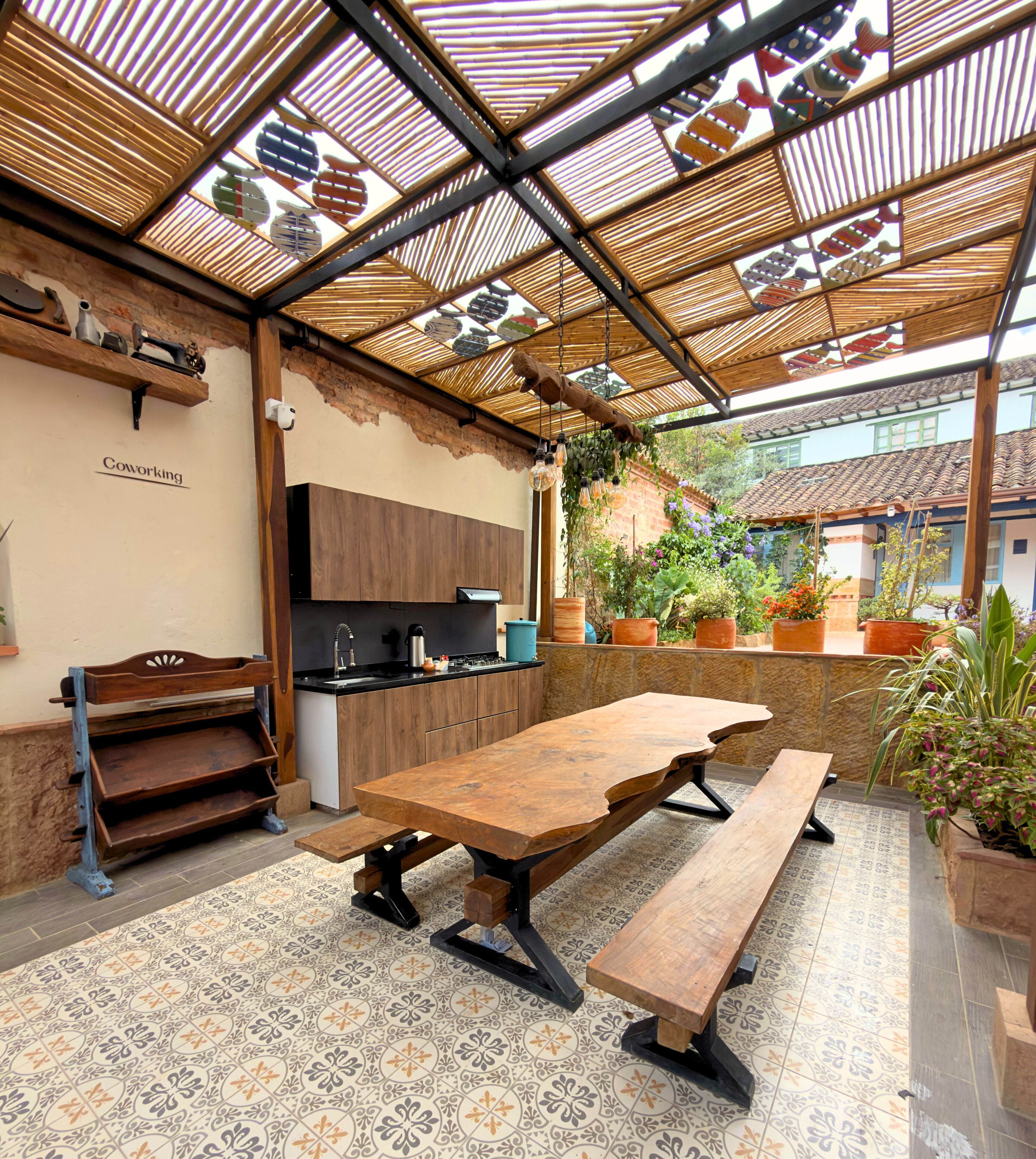 Un espacio comedor al aire libre cubierto, con una mesa y bancos de madera, una zona de cocina con encimera negra y plantas en macetas.