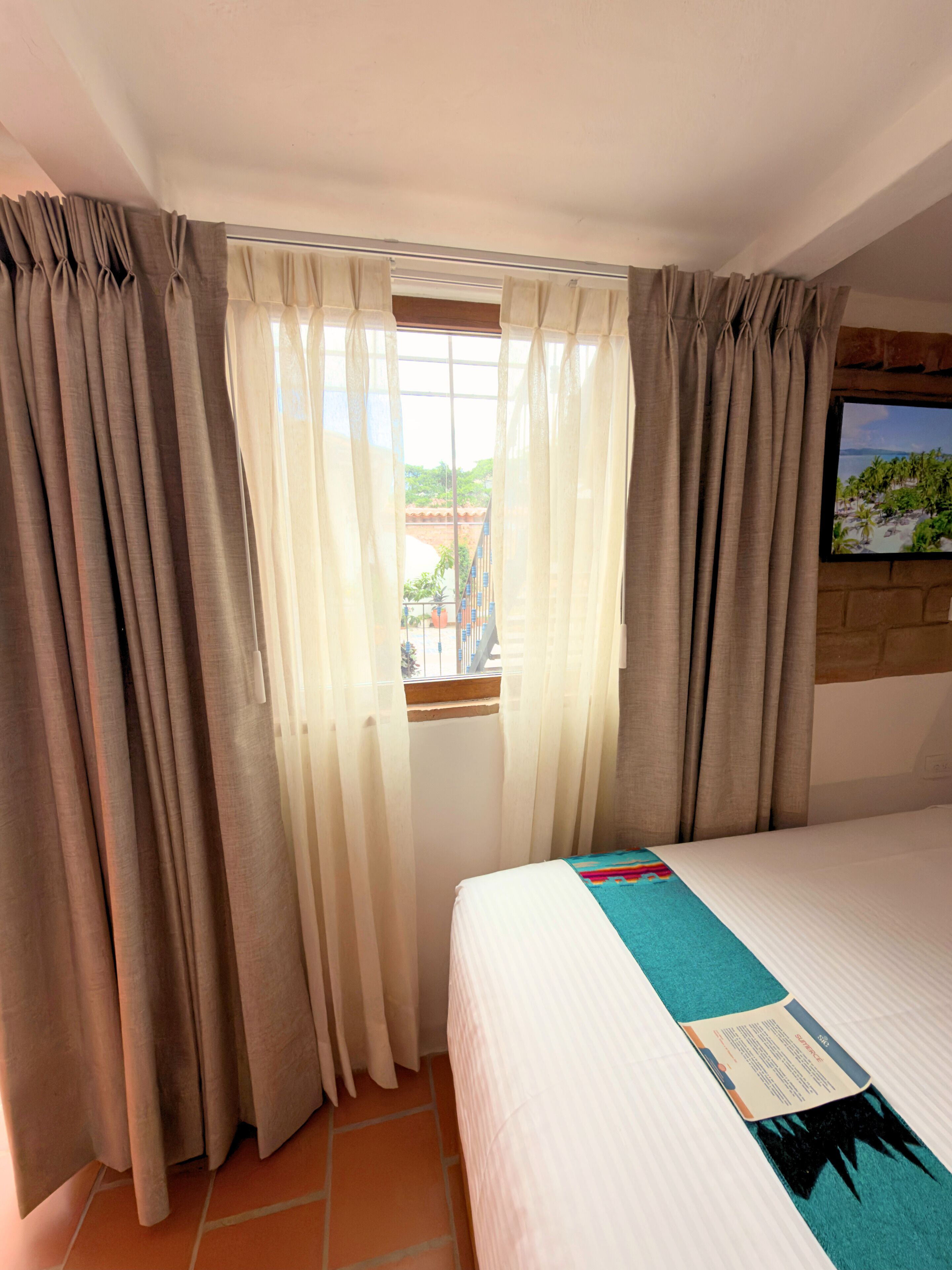 A hotel room with a bed, curtains, a window, and a television on the wall.