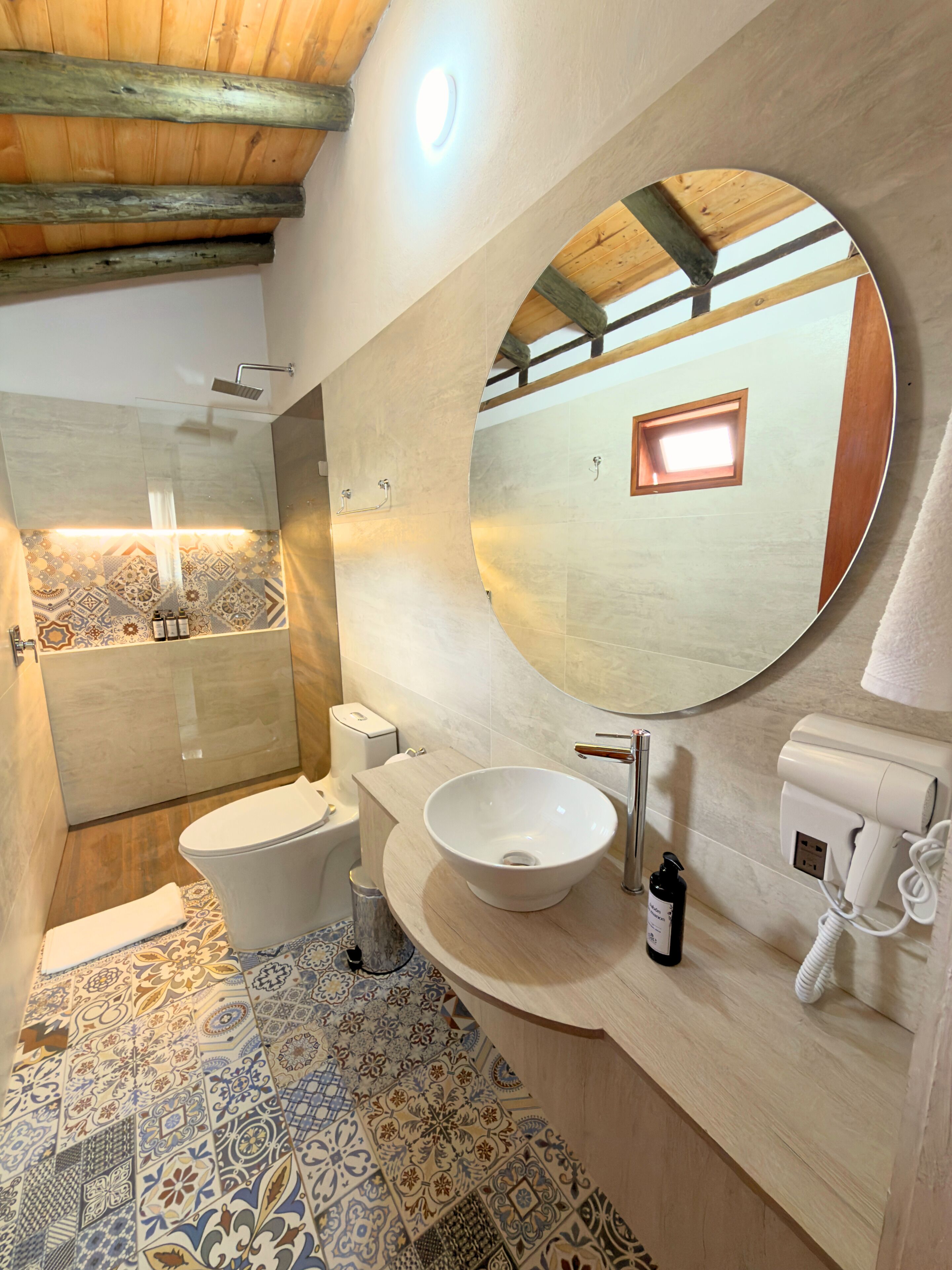 Un bany amb sostre de fusta, un mirall rodó gran, un lavabo, un vàter i una barra de tovalloles.