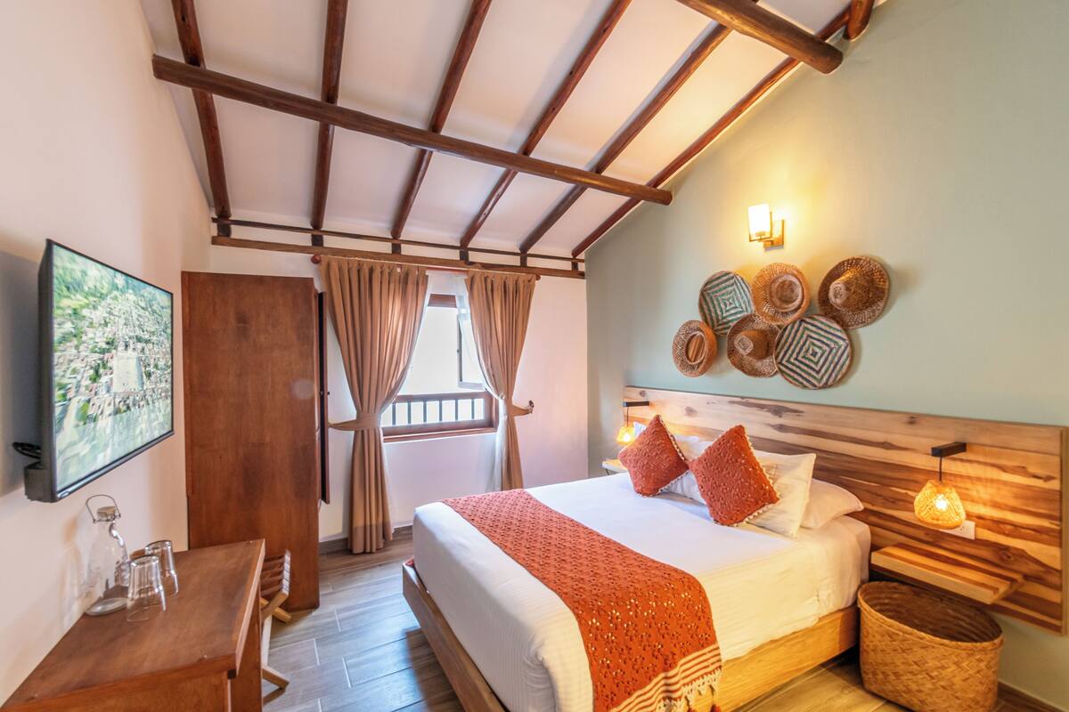 A bedroom with a wooden bed, a TV mounted on the wall, a wooden table with glasses, and a window with curtains.