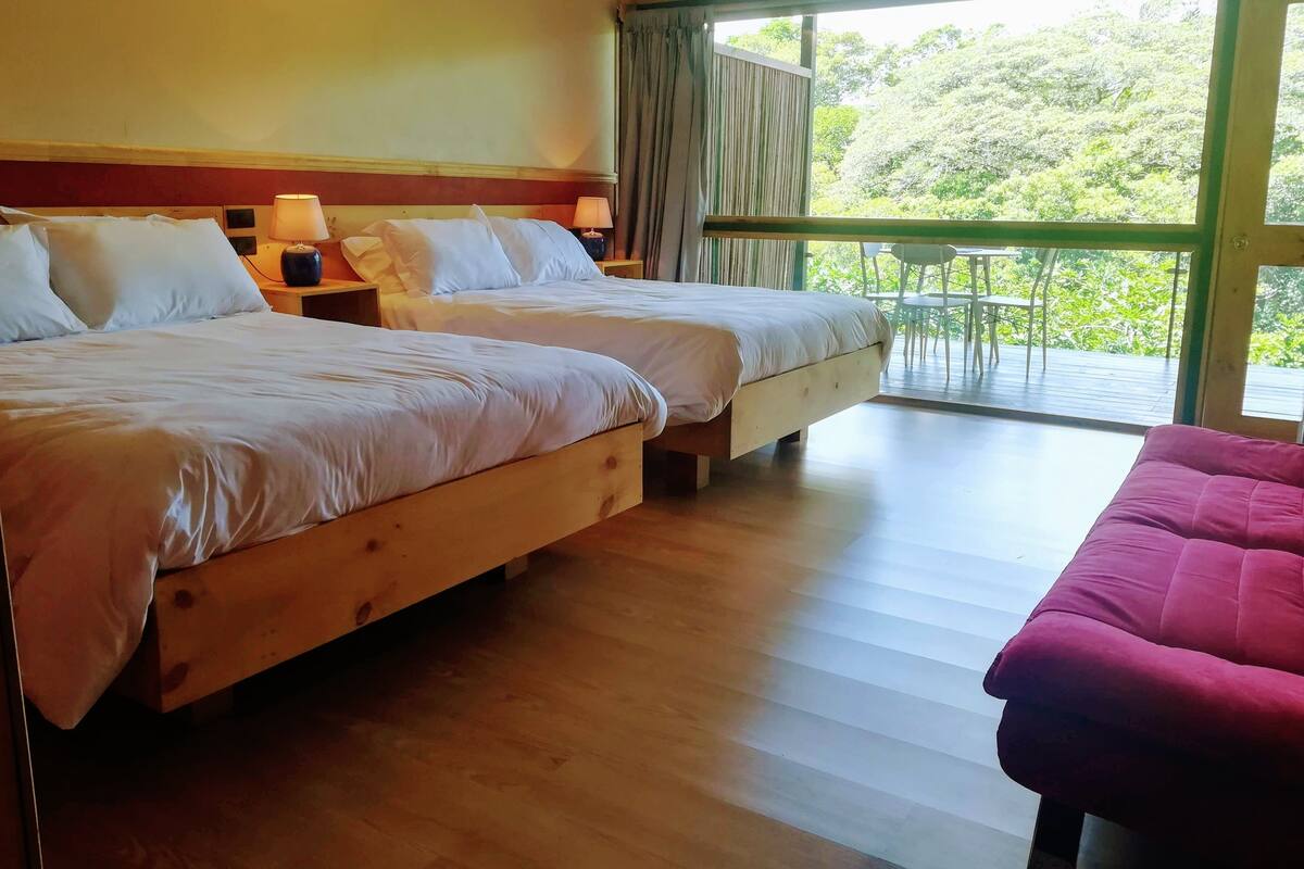A room with two wooden beds, a wooden bench, and a balcony with a view of greenery.
