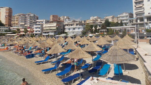 Una spiaggia con file di lettini e ombrelloni di paglia, cielo sereno e edifici sullo sfondo.