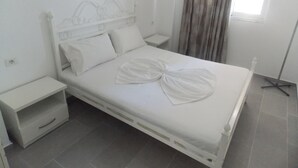 Un letto rifatto con lenzuola e cuscini bianchi, un comodino e una lampada da terra in una stanza con pavimento piastrellato di grigio.