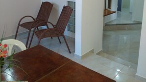 A dining area with a wooden table and wicker chairs, a painting on the wall, and a view into a hallway with a tiled floor. - Residencial Praia Bombas (Bombinhas)