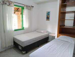 Una habitación pequeña y limpia con dos camas, una ventana con cortinas, un cuadro en la pared y un estante de madera.