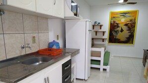 A kitchen with white cabinets, a stainless steel sink, and a refrigerator. There is a painting on the wall and a small shelf with decorative items. - Residencial Praia Bombas (Bombinhas)